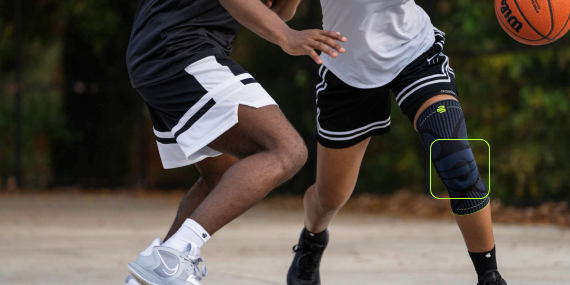 Due giocatori di basket su un campo esterno in piena azione. Il giocatore in primo piano indossa una ginocchiera nera e palleggia mentre viene marcato. La scena trasmette energia e reattività.
