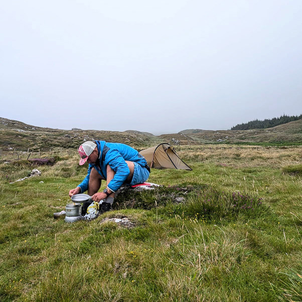 Sfida fitness: Quando la natura diventa la tua cucina e ogni pasto è un'avventura. Una persona con una giacca blu è seduta su un prato in un paesaggio collinare davanti a una piccola tenda, mentre prepara un pasto su un fornello da campeggio. Il cielo è nuvoloso e il paesaggio appare selvaggio e incontaminato.