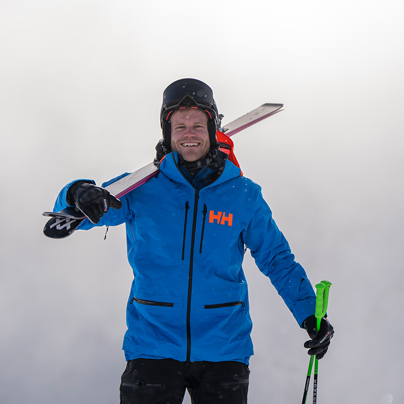 Pronti per la pista Un uomo in piedi davanti a uno sfondo nebbioso, indossa una giacca blu e un casco con occhiali da sci e ha gli sci in spalla.
