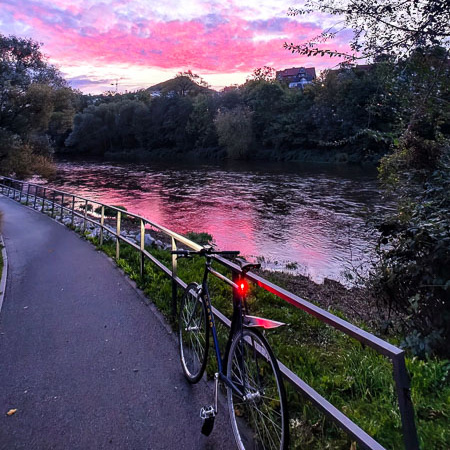 Incanto al tramonto – quando il percorso fitness diventa un paesaggio romantico. Una bicicletta appoggiata a una ringhiera lungo il fiume, mentre il cielo si tinge di sfumature viola e rosa. Sullo sfondo, i colori del cielo si riflettono nell'acqua calma del fiume.