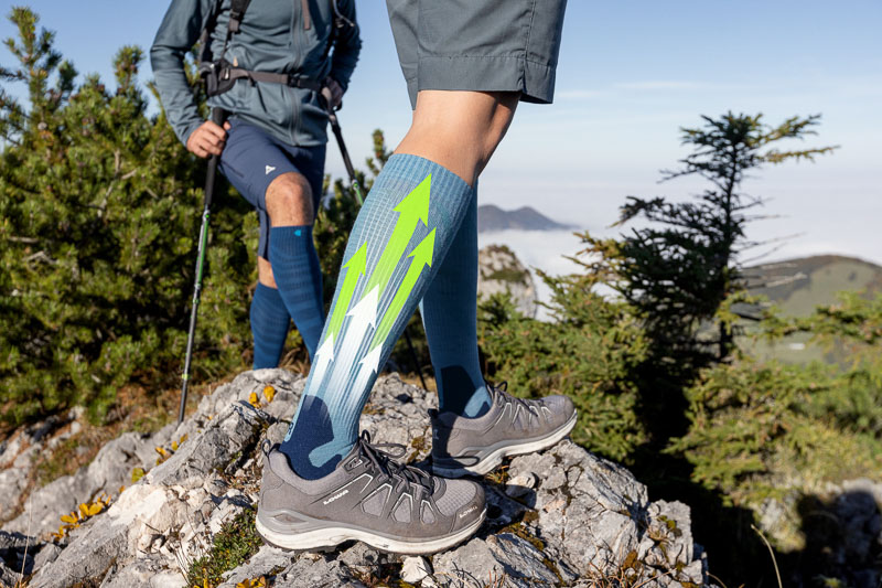 Due escursionisti su un sentiero roccioso di montagna indossano calze a compressione con un design dinamico caratterizzato da frecce verdi e bianche. Le calze evidenziano supporto e flusso energetico durante l'attività.