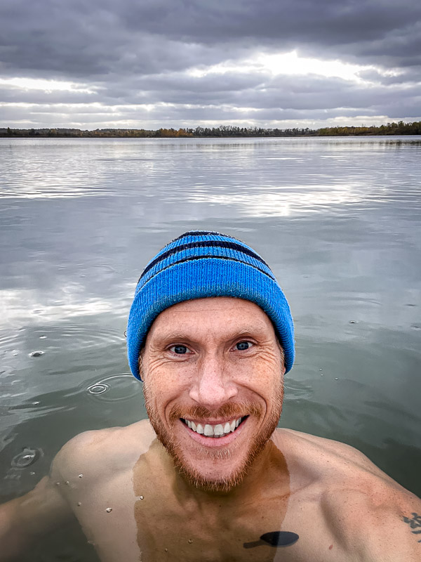 Un sorriso tra i brividi – il tuffo definitivo nell’acqua gelata! Un uomo con un berretto blu sorride alla fotocamera mentre si trova in un lago freddo. Il cielo è nuvoloso e l'acqua calma riflette le nuvole grigie.