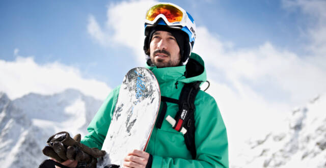 Snowboarder con una giacca verde con tavola in mano davanti a uno sfondo di montagna