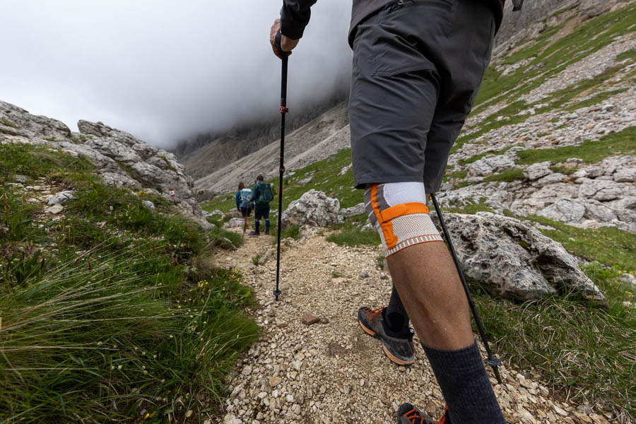 Un escursionista con bastoncini da trekking e ginocchiera cammina su un sentiero roccioso attraverso un paesaggio montano. Sullo sfondo si vedono altri escursionisti e nuvole sopra le montagne.