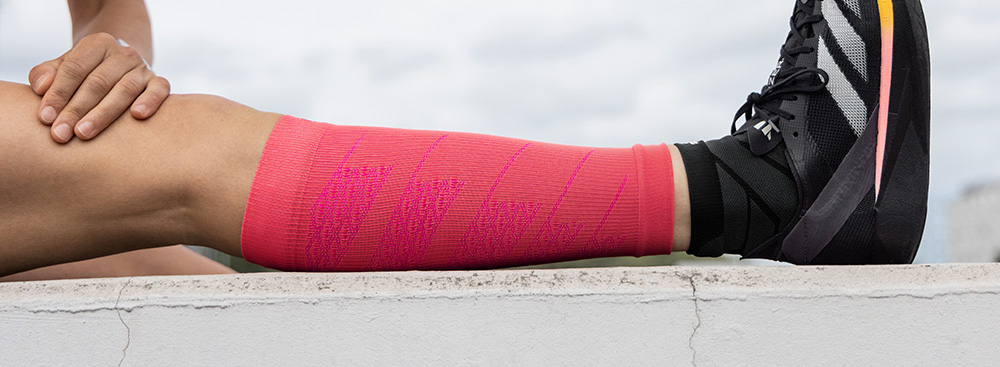 Power in pink – pronti a correre! Un atleta è seduto a terra e allunga la gamba tesa. Indossa manicotti a compressione rosa acceso sul polpaccio, abbinati a scarpe sportive nere. L’immagine mette in risalto la vestibilità aderente e il supporto durante il riscaldamento.