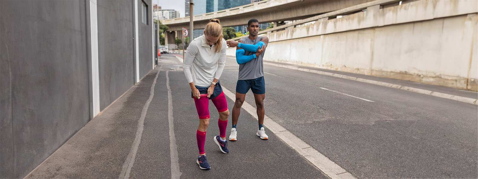 Guarda anche bene per la pausa: nessun problema con i nostri prodotti eleganti Una donna con maniche a compressione rosa e un uomo con maniche a braccio blu e calze a corto di corsa si fa una pausa sul ciglio della strada di una grande città e si allunga