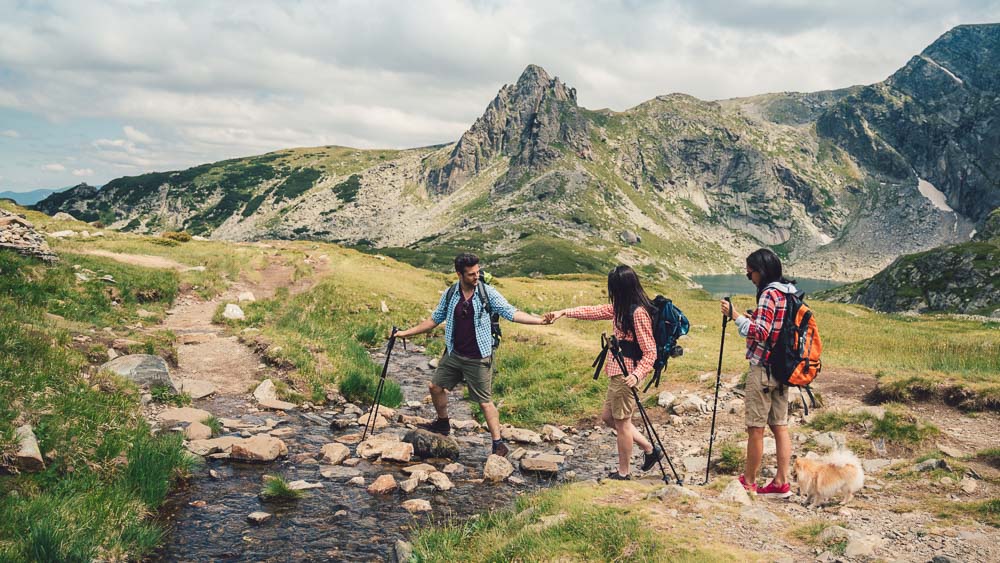 Insieme nel paesaggio alpino Tre escursionisti, due donne e un uomo, attraversano insieme un ruscello alpino con cime rocciose sullo sfondo. L’uomo tende la mano per aiutare – al centro ci sono il gioco di squadra e il contatto con la natura.