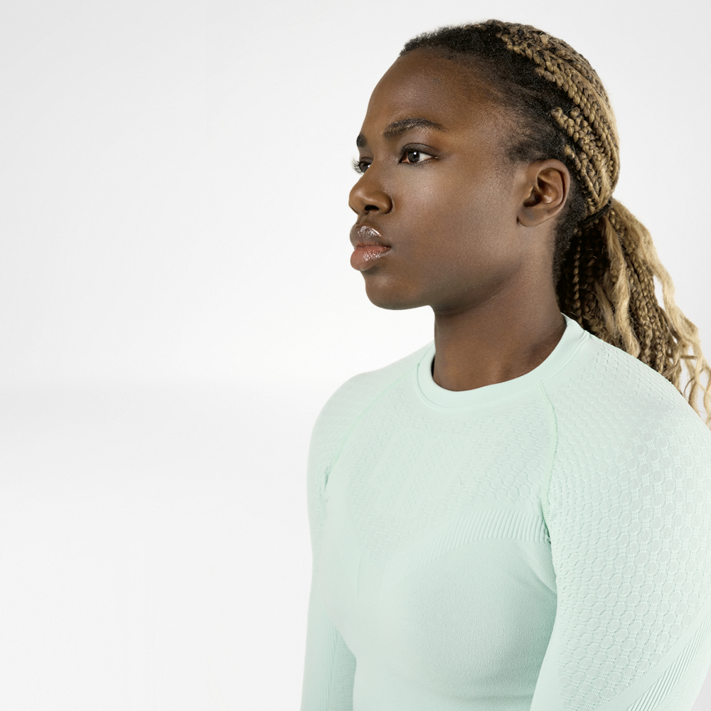 Una donna sportiva con i capelli intrecciati che indossa una maglia a compressione verde chiaro a maniche lunghe vista di lato. Ha un'espressione concentrata e determinata sul volto.