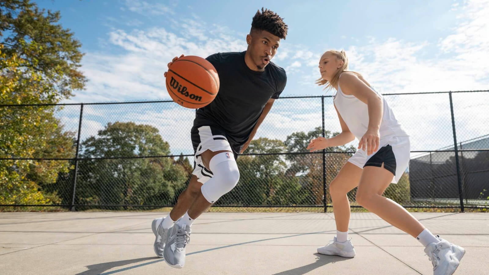 Un giocatore di basket avanza rapidamente su un campo all’aperto con la palla, seguito da un avversario. Indossa un tutore bianco al ginocchio per una maggiore stabilità. Intorno si vedono alberi autunnali e recinzioni.