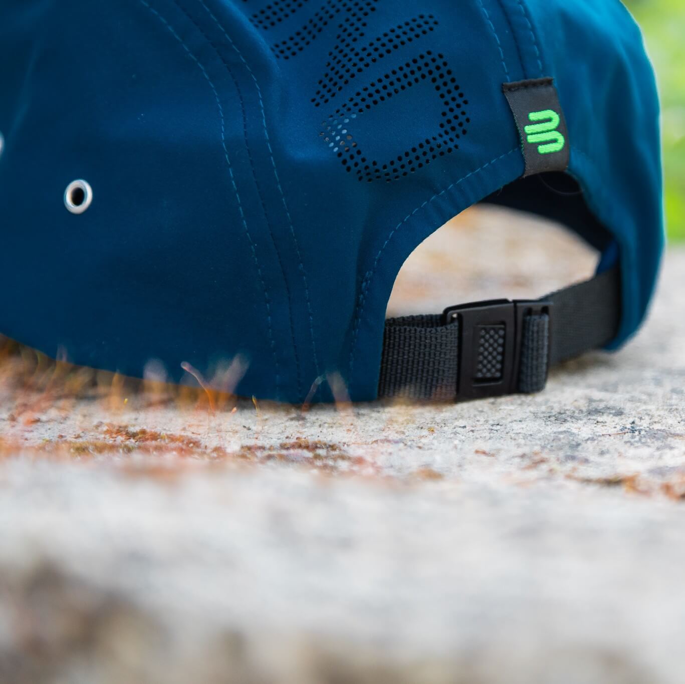 Primo piano del retro di un cappellino blu Bauerfeind Sports con chiusura nera regolabile, etichetta con logo verde, occhiello metallico laterale e scritta traforata – dettagli pratici per una vestibilità stabile durante le attività outdoor.