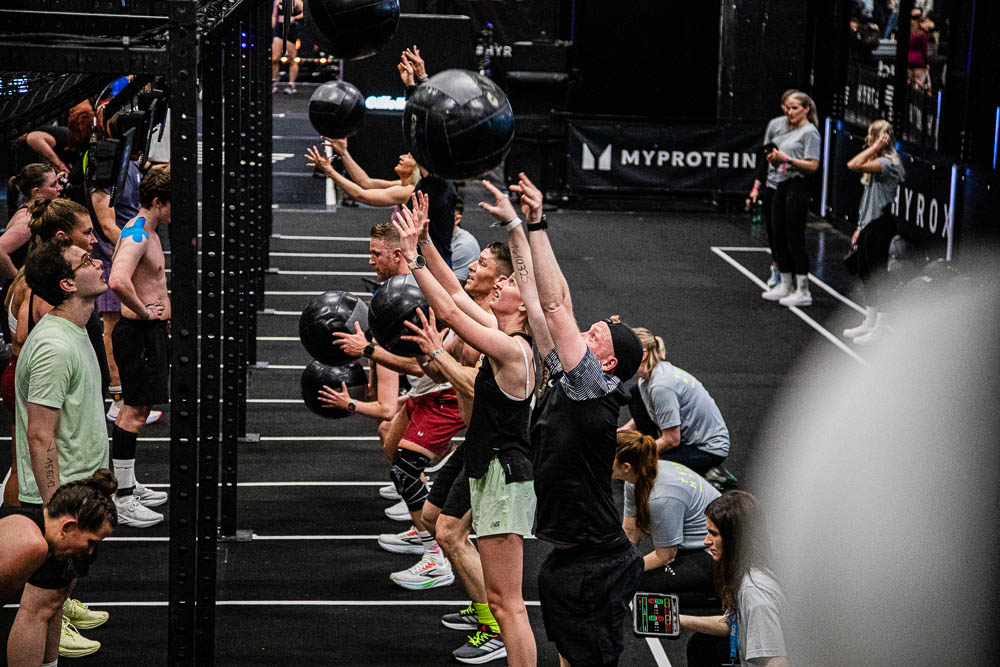 Atleti impegnati nei wall ball in una zona workout HYROX, mentre lanciano medicine ball nere verso l’alto e giudici e staff seguono le ripetizioni; la scena mostra intenso lavoro full body, concentrazione e un’atmosfera di gara motivante.