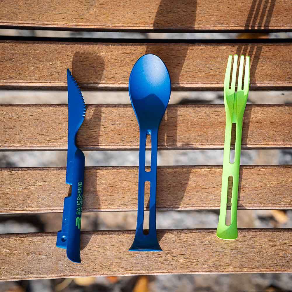 Il set completo: Coltello, forchetta e cucchiaio per chi è in movimento Un set di posate smontato con un coltello blu, un cucchiaio blu e una forchetta verde sono affiancati su una panchina di legno.