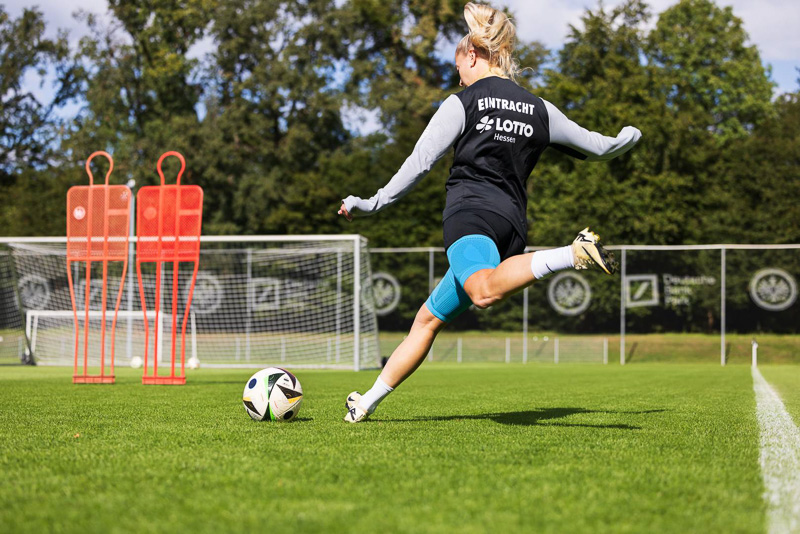 Giocatrice dell'Eintracht Francoforte durante un allenamento di tiro Una giocatrice dell'Eintracht Francoforte tira con forza verso la porta sullo sfondo. Indossa abbigliamento da allenamento con il logo Eintracht e una ginocchiera blu di Bauerfeind Sports.