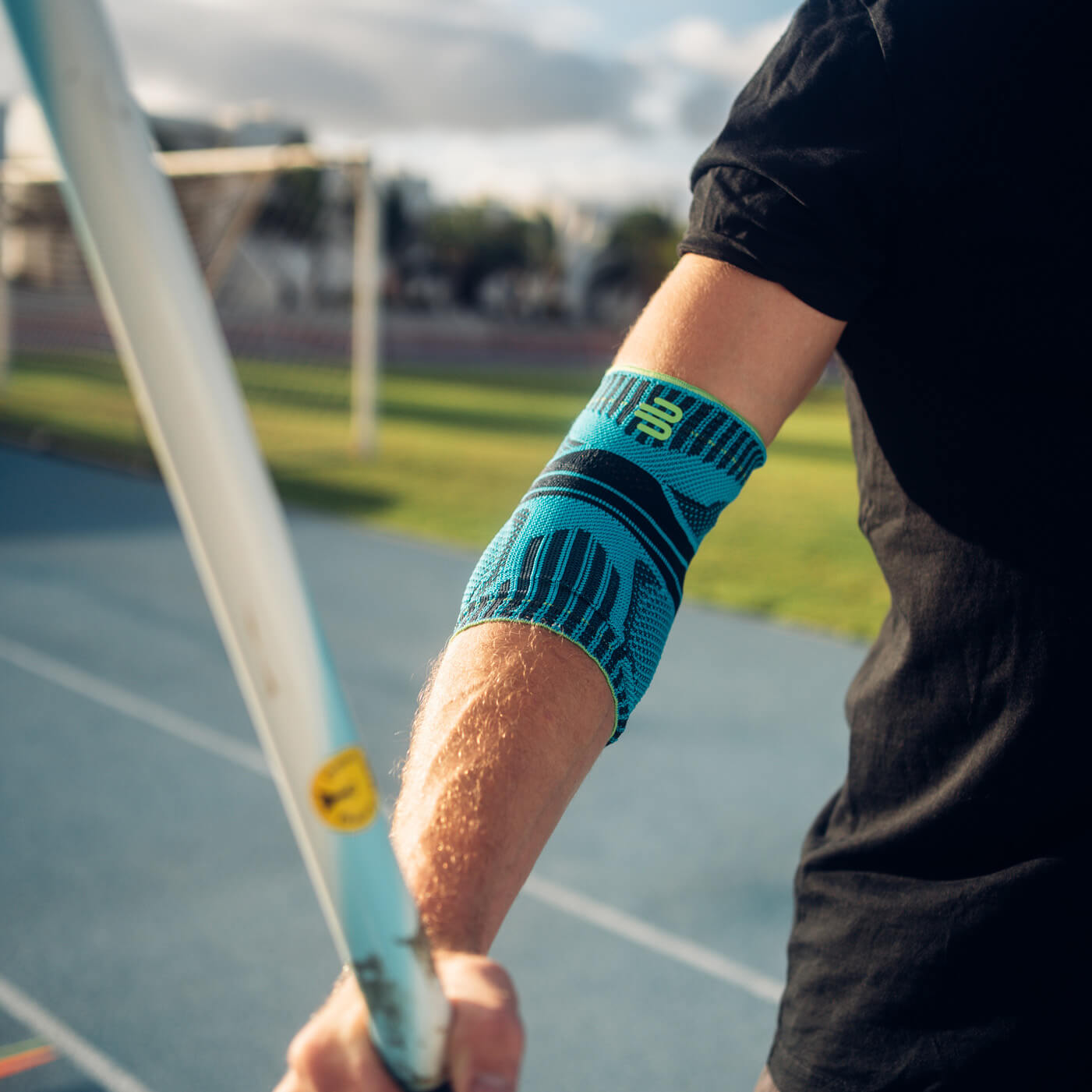Precisione e potenza – con supporto stabile per il tuo gomito. Primo piano di un braccio con una fascia per il gomito blu durante un lancio del giavellotto. Sullo sfondo si vedono una pista di atletica e un paesaggio soleggiato.