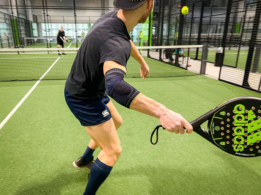 Massima concentrazione nella partita di padel! Un giocatore di padel in maglietta e pantaloncini neri colpisce la palla con massima concentrazione. Indossa una fascia sportiva blu scuro sul braccio destro per maggiore supporto.