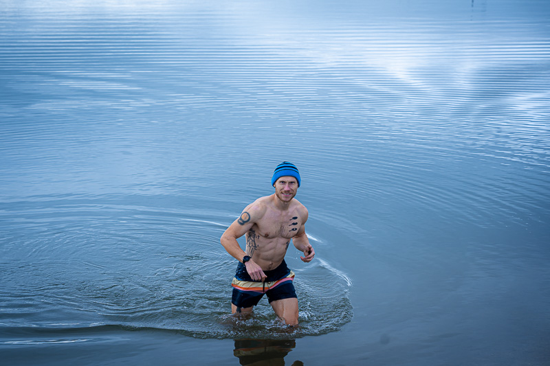 Un uomo esce dal lago, ripreso mentre fa un bagno di ghiaccio in costume da bagno e con berretto blu nell'ambientazione autunnale.