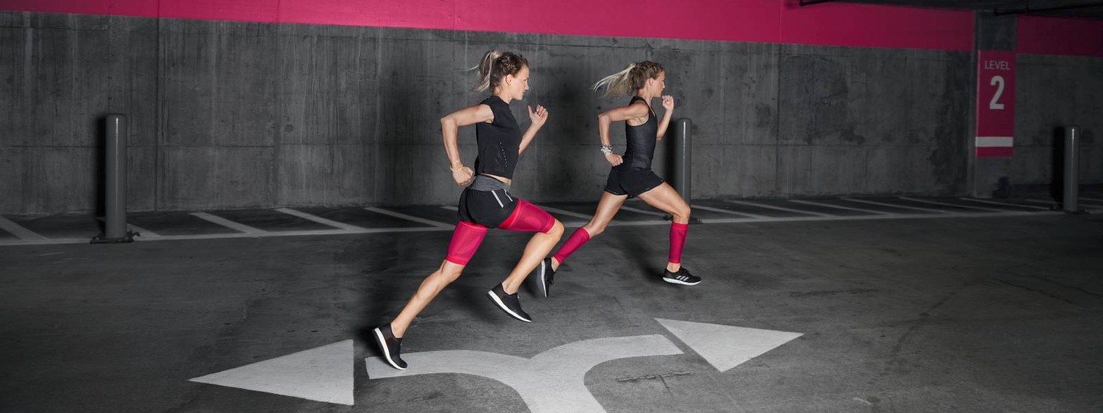 Accento di colore forte per muscoli potenti: le nostre maniche a compressione rosa Due corridori con maniche a compressione rosa corrono attraverso un garage
