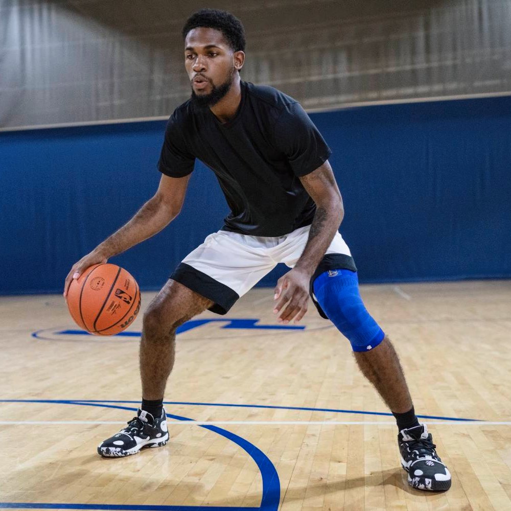 Un giocatore di basket in posizione difensiva su un campo indoor, indossa una ginocchiera a compressione blu sulla gamba destra. Il pavimento in legno riflette la luce della palestra.