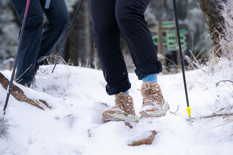 Passo dopo passo nella neve – Ogni impronta conta! Primo piano di scarponi da trekking che calpestano la neve profonda. I piedi appartengono a escursionisti con pantaloni neri e calzini blu. I bastoncini con punte gialle affondano nella neve, mentre il terreno è coperto di neve e rocce.