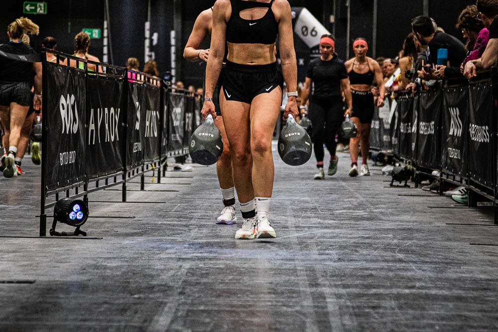 Atleta che trasporta due kettlebell pesanti lungo il percorso HYROX durante una competizione, mentre altri partecipanti la seguono e il pubblico osserva ai lati della corsia;
