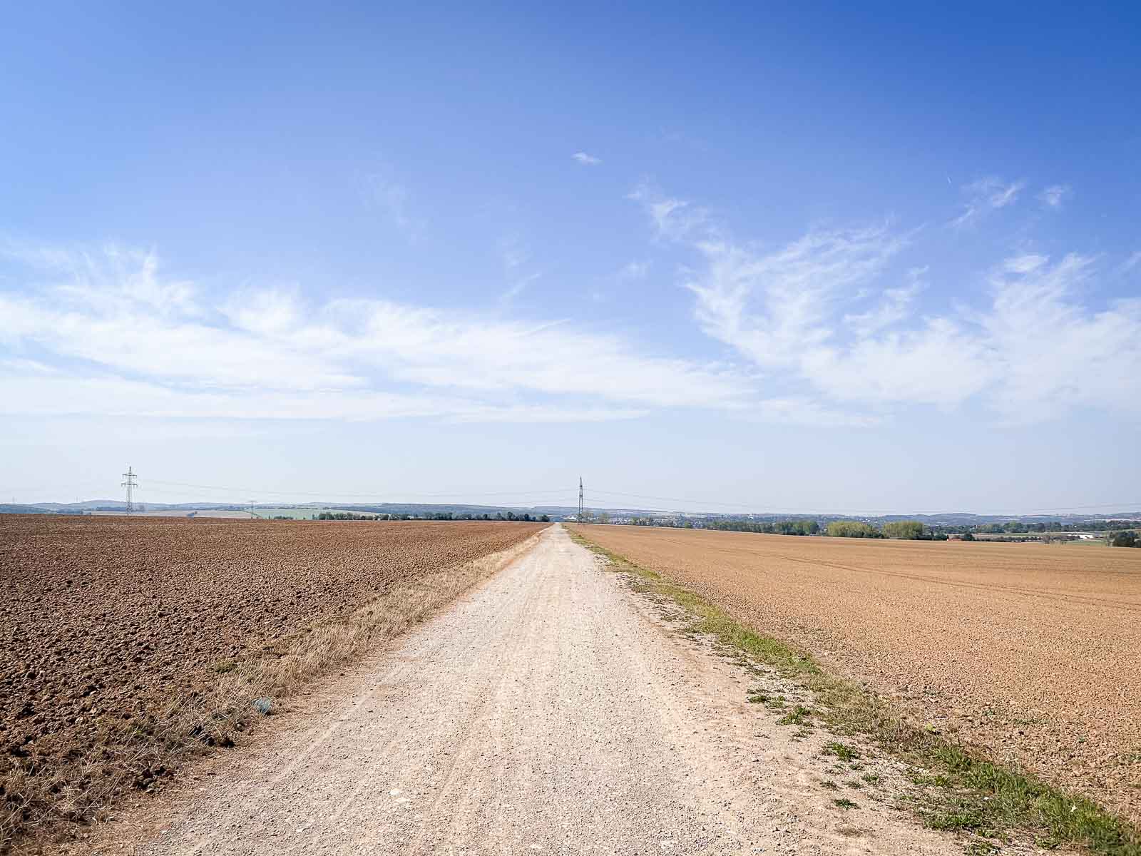Un lungo sentiero di ghiaia rettilineo che attraversa un vasto paesaggio con cieli sereni e campi su entrambi i lati. Percorso di corsa ideale per corse di resistenza.