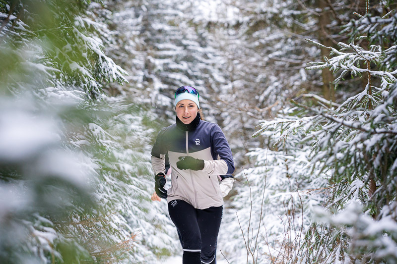 Corsa invernale: Tra abeti innevati sul sentiero della determinazione. Una runner corre lungo un sentiero innevato in un bosco. Gli alberi intorno sono coperti di neve fresca. Indossa una giacca da corsa grigio chiaro con accenti neri, pantaloni neri e una fascia blu con occhiali da sole.
