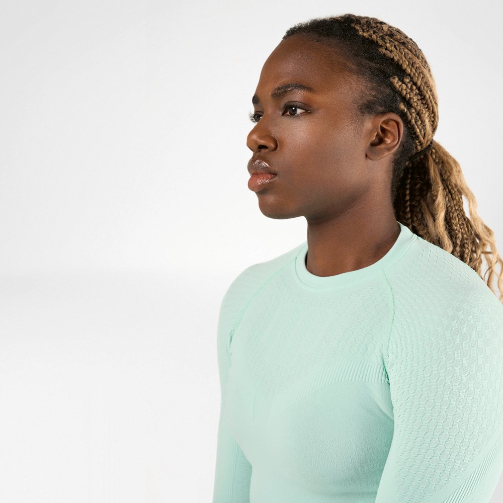 Una donna sportiva con i capelli intrecciati che indossa una maglia a compressione verde chiaro a maniche lunghe vista di lato. Ha un'espressione concentrata e determinata sul volto.