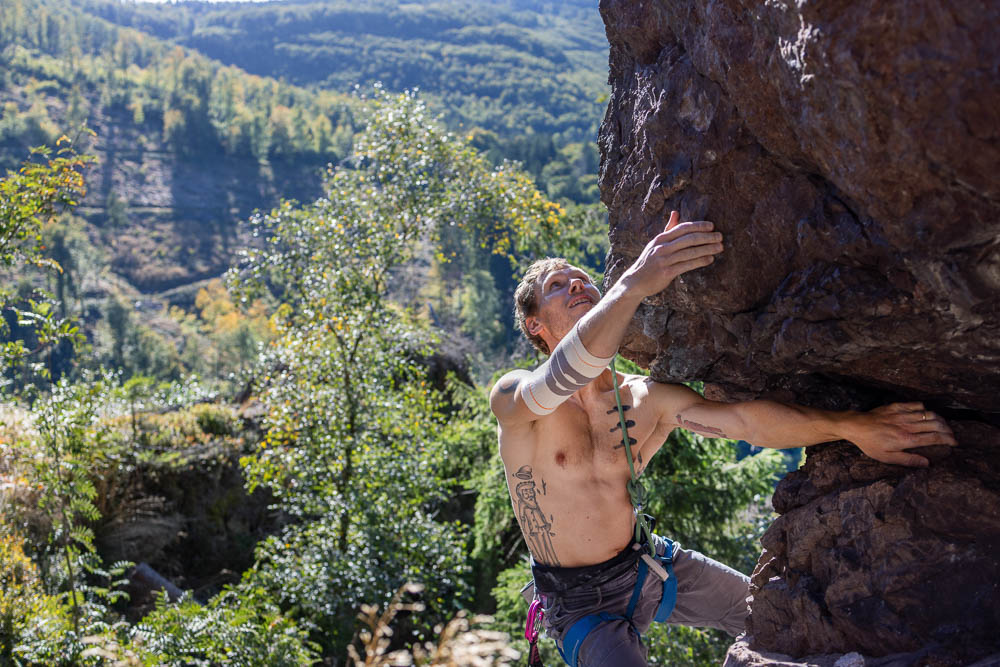 Inquadratura ampia di un climber su un roccione a strapiombo con vista sulla valle; indossa il Bauerfeind Sports Outdoor Compression Elbow Support color sandstone mentre si sostiene sulla roccia.
