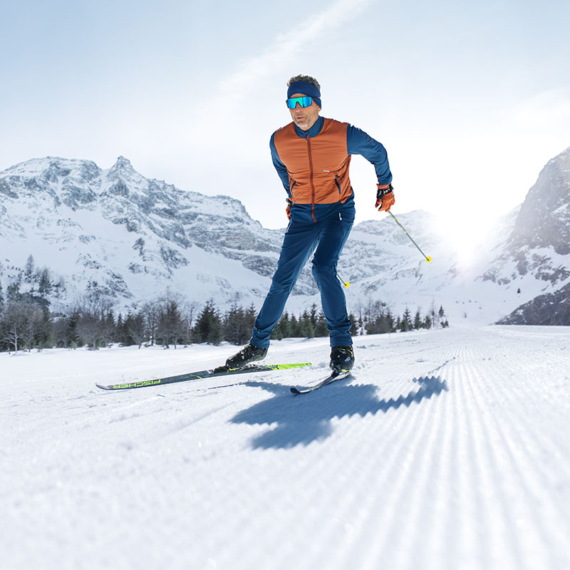 Avventura in pista: Sciatori in look invernale Un uomo con occhiali da sci blu, fotografato frontalmente, sta sciando in un paesaggio invernale. Indossa una giacca arancione e pantaloni da sci blu.