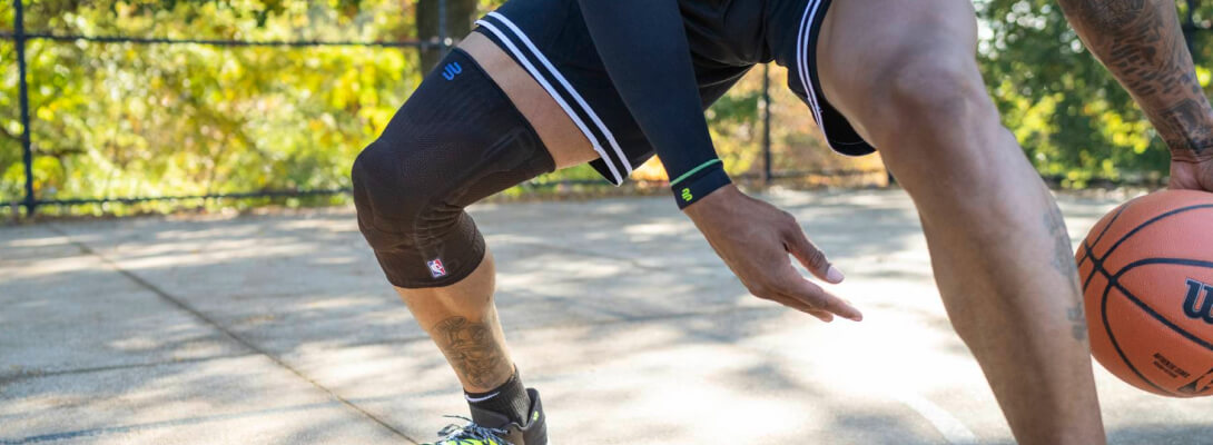 Giocatore di basket con ginocchiera nera in palleggio Un giocatore di basket indossa un tutore per il ginocchio nero a compressione mentre palleggia su un campo esterno. Il tutore mostra chiaramente il logo NBA e quello del produttore.