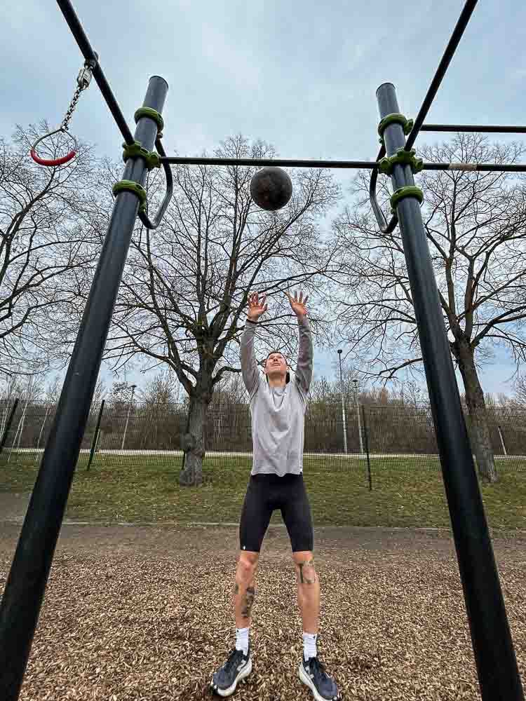 Un atleta lancia una palla medica verso l’alto in un parco outdoor sotto una struttura da calisthenics. La scena mostra un esercizio funzionale di forza e conditioning adatto all’allenamento HYROX senza palestra.