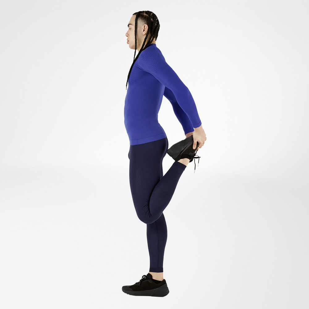 Un atleta indossa un abito a compressione blu, composto da una maglia a maniche lunghe e collant, esegue un esercizio di stretching, tenendo una gamba indietro, su uno sfondo bianco. L'abito offre un alto comfort e un supporto muscolare ottimale.