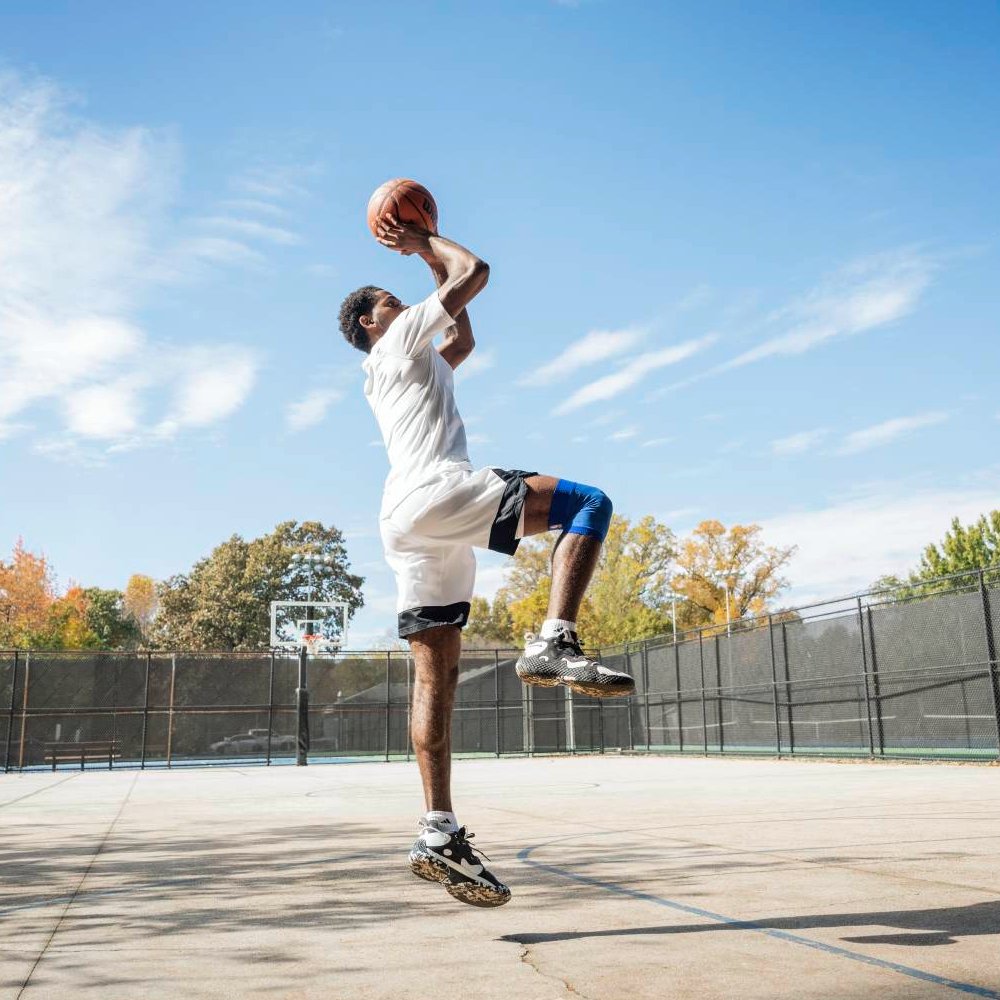 Un giocatore di basket con un supporto per il ginocchio blu dei Dallas Mavericks mentre esegue un tiro in salto su un campo all'aperto. Il supporto aiuta il ginocchio durante il movimento.