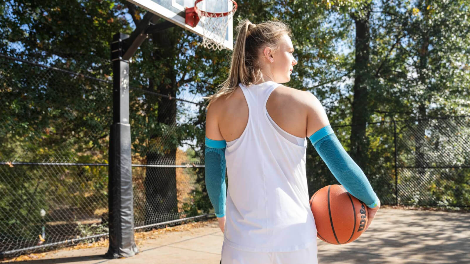 Pronta per il prossimo tiro. Una giocatrice di basket si trova su un campo all’aperto con un pallone da basket nella mano destra. Indossa manicotti compressivi azzurri e guarda concentrata verso il canestro.