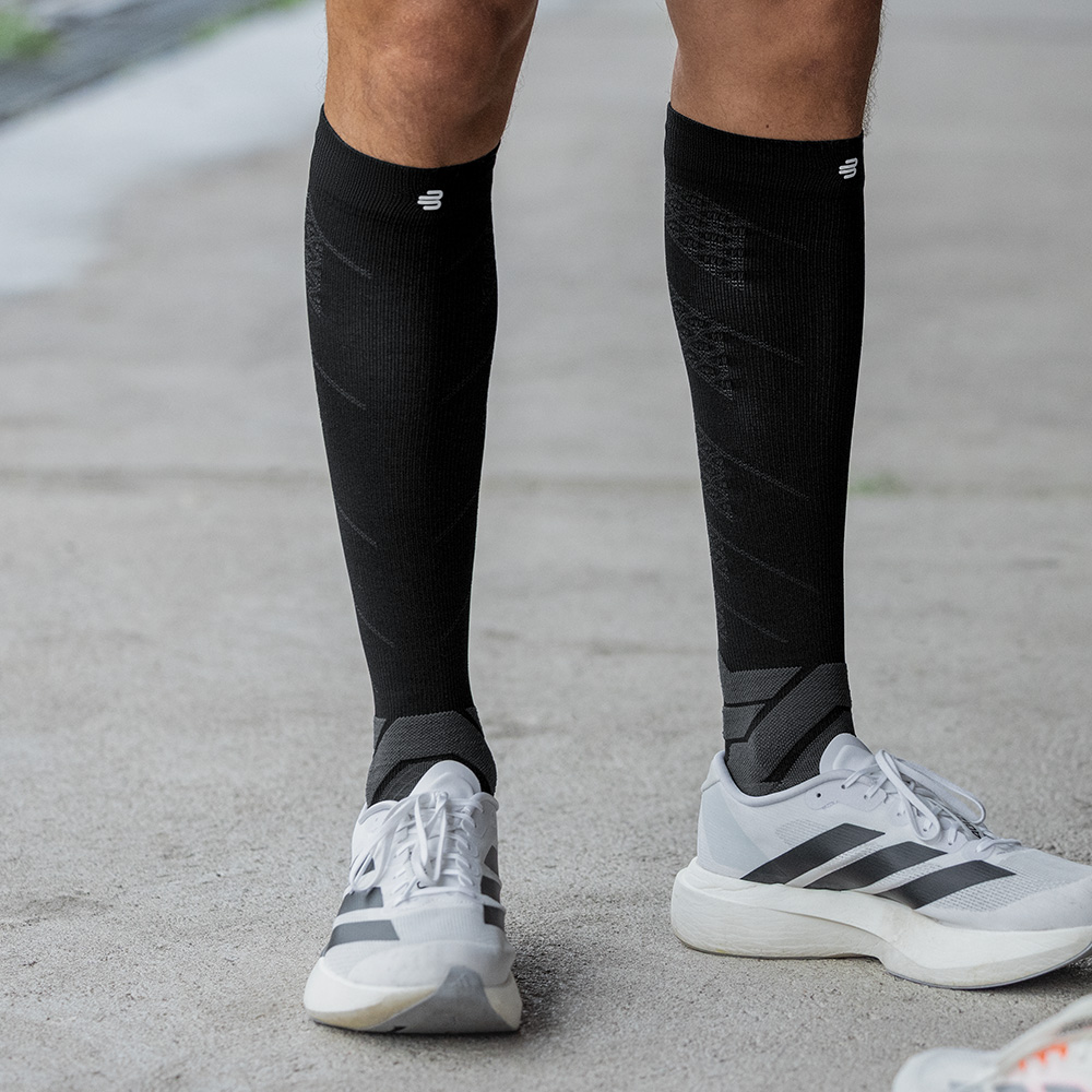 Un uomo sportivo è in piedi frontalmente su asfalto, indossando calze a compressione nere Bauerfeind e scarpe da corsa bianche.