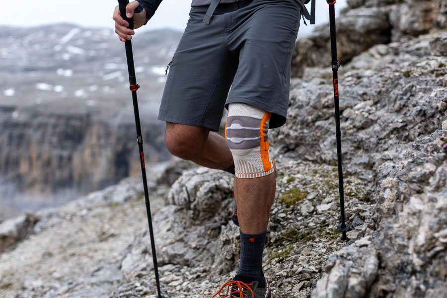 Un escursionista con bastoncini da trekking e ginocchiera arancione cammina su un sentiero montano roccioso. Sullo sfondo si vedono scogliere frastagliate e paesaggi montani nebbiosi.