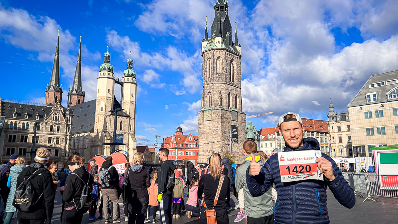 Partecipante al punto di partenza della Mitteldeutscher Halbmarathon sulla piazza del mercato di Halle (Saale) con la Marktkirche e la Torre Rossa sullo sfondo. Un partecipante alla Mitteldeutscher Halbmarathon alza il suo numero di pettorale nella piazza del mercato di Halle (Saale). Dietro di lui si vedono la Torre Rossa e la Marktkirche. In primo piano, corridori e spettatori si preparano alla gara.