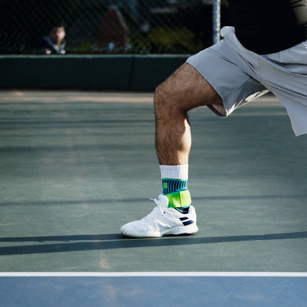 Un tennista su un campo in cemento, in movimento durante il servizio. Indossa la Bauerfeind Sports Ankle Support Rivera in blu-verde sopra i suoi calzini sportivi bianchi.
