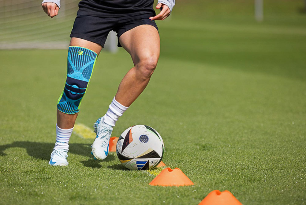 Una calciatrice si allena sul campo indossando un tutore per ginocchio Bauerfeind. L'attenzione è sulla stabilità e sul supporto forniti dal tutore durante i movimenti dinamici, come il dribbling intorno ai coni.