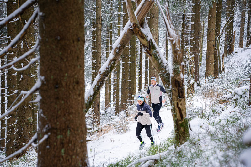 Corsa invernale sotto l'arco della natura Due corridori corrono attraverso una foresta innevata. Il sentiero è circondato da alti abeti coperti di neve, mentre un arco naturale di rami attraversa il percorso. Entrambi indossano abbigliamento invernale da corsa e fasce per la testa.