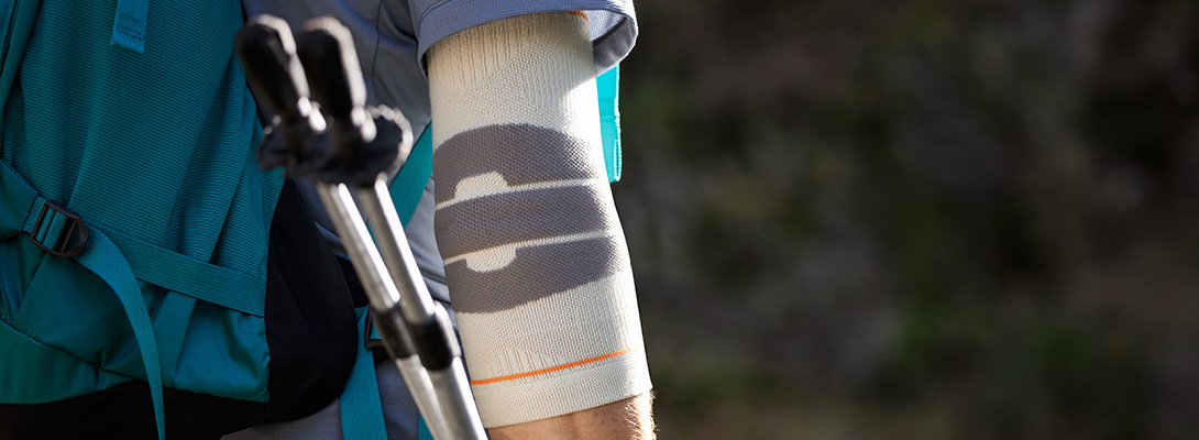 Un escursionista con zaino utilizza bastoncini da trekking su un sentiero stretto. Al gomito indossa un Outdoor Compression Elbow Support che offre sostegno mirato e stabilità durante le escursioni più impegnative.