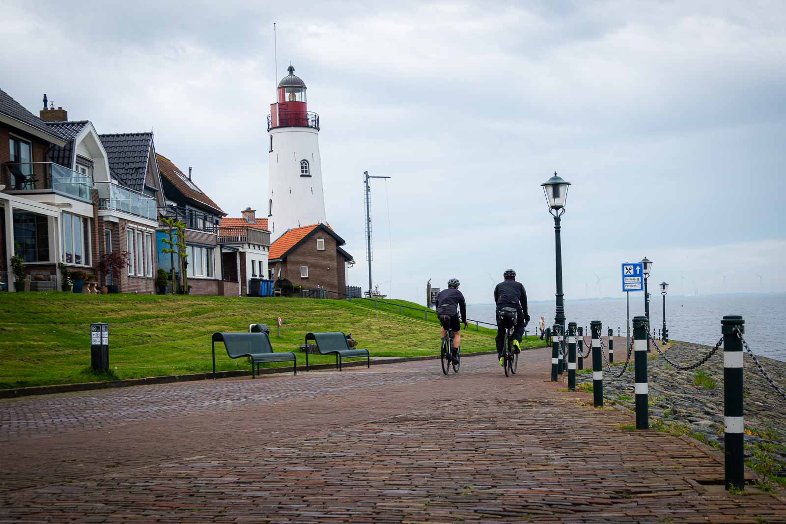 Ciclisti ultraciclisti su un lungomare con un faro Due ciclisti ultraciclisti di spalle che percorrono un lungomare dove sorge un faro.