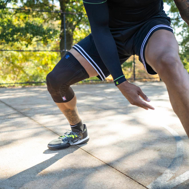 Un giocatore di basket in movimento su un campo all’aperto, indossa una manica a compressione nera e un supporto per il ginocchio nero con logo NBA.