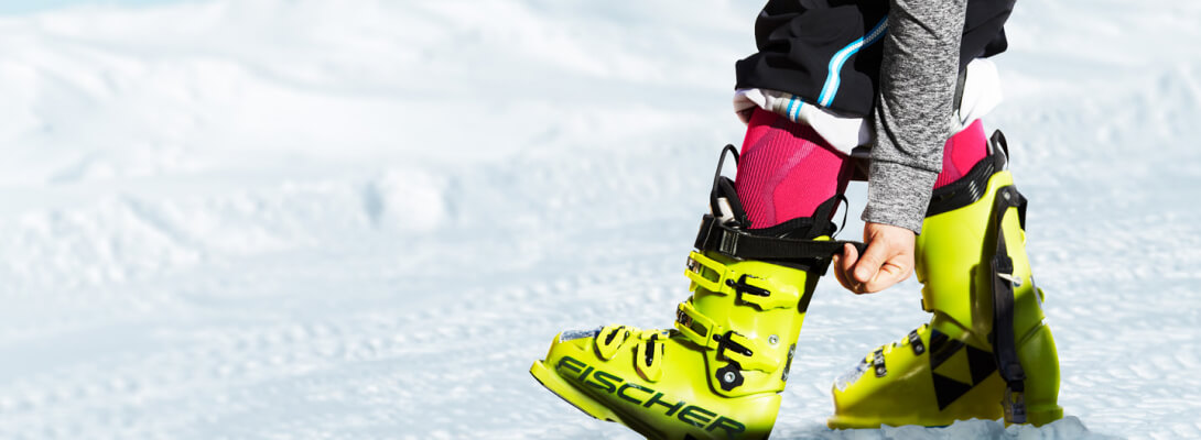 Dettaglio dell'immagine che mostra una persona con gli scarponi da sci e i calzini da sci sotto, che afferra la chiusura degli scarponi