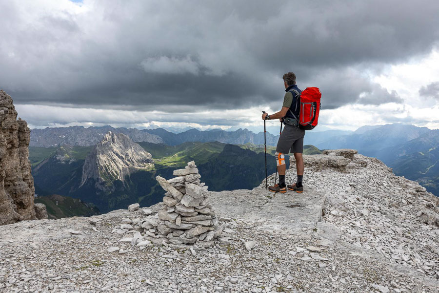 Escursionisti su un promontorio roccioso con una ginocchiera da trekking Una persona con uno zaino rosso è in piedi su un affioramento roccioso nelle Dolomiti, guardando lontano. Il cielo è nuvoloso e c'è una costruzione di ometti di pietra in primo piano.