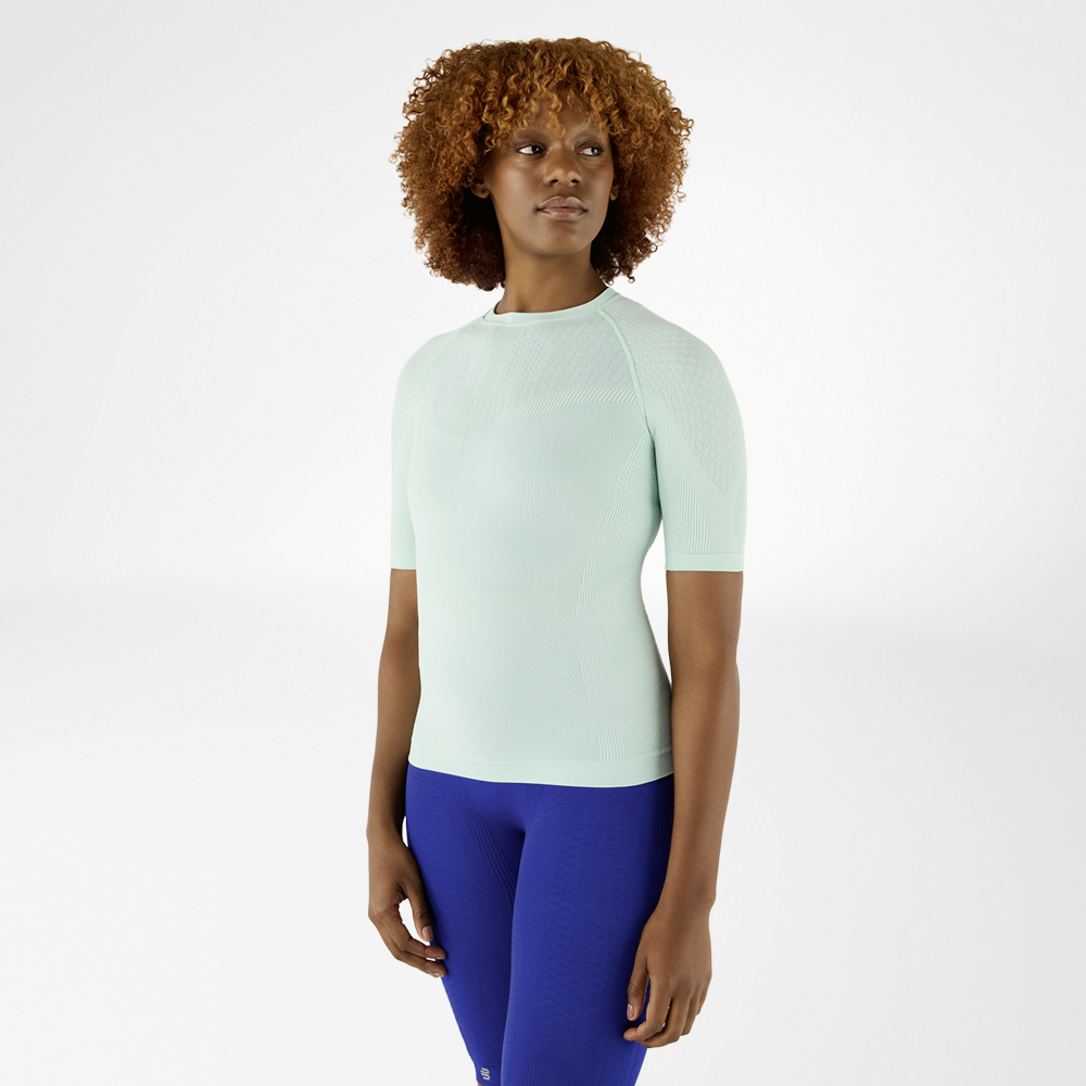 Donna con maglia a compressione verde menta e leggings blu, in piedi rivolta in avanti.