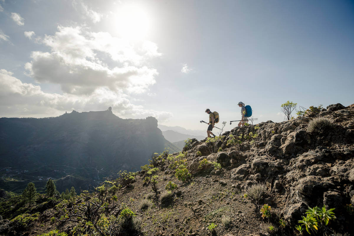 Largo angolo: due escursionisti con grandi zaini e bastoncini vanno con cura una catena montuosa con pietre e piccoli arbusti verso il basso sullo sfondo si possono vedere altre montagne