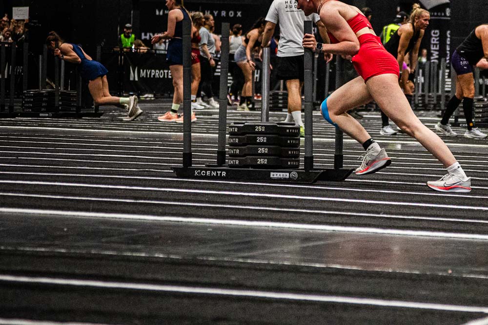 Atleta con outfit sportivo rosso che spinge una slitta pesante su un’area indoor segnata durante una competizione HYROX, mentre altri partecipanti lavorano alle proprie stazioni sullo sfondo