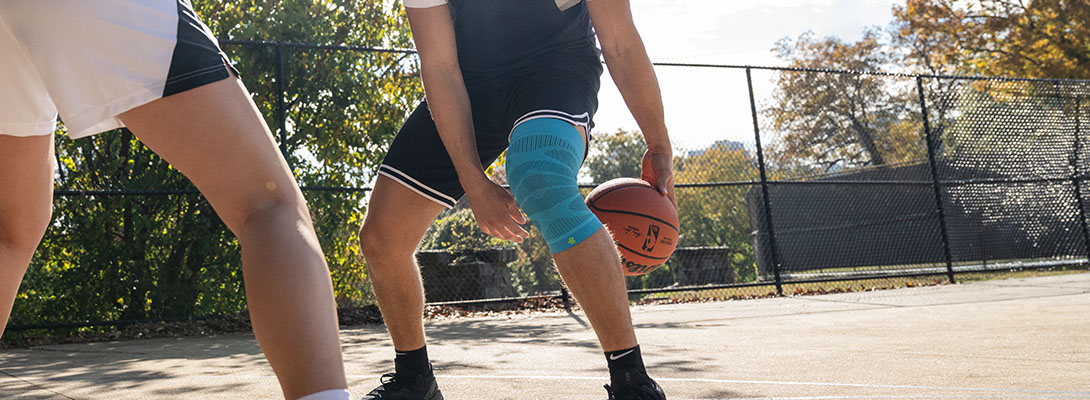 Manica al ginocchio al basket Un giocatore di pallacanestro si trova su un campo in cemento armato circondato da una recinzione ed esegue un trucco con la palla facendola passare attraverso le sue due gambe. Un altro giocatore è in piedi di fronte a lui. Indossa una ginocchiera.