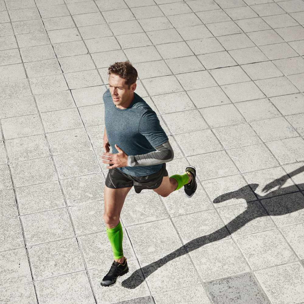 Un corridore atletico con maglia blu e pantaloncini neri sprinta su una superficie urbana lastricata. Indossa manicotti a compressione verdi per migliorare la circolazione e sostenere i muscoli. La sua postura trasmette energia e dinamismo.